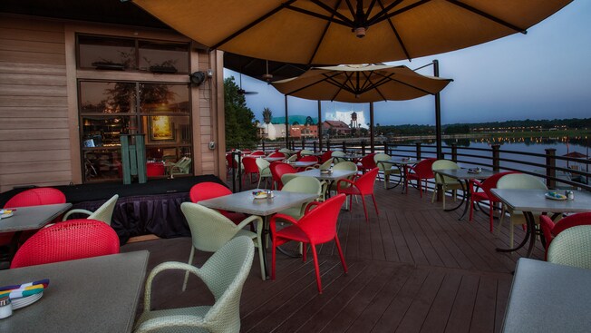 On the lakeside deck, large umbrellas hang above tables with red and white wicker chairs 