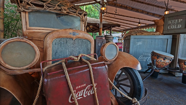 Antique car with netting on its roof, Coca-Cola® cooler tied to its front and drums in the background