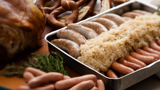 Pan filled with sauerkraut and 2 types of German sausages sitting on a table next to meats and pretzels