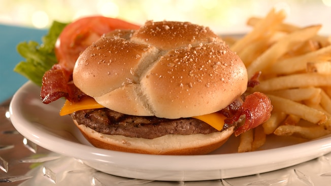 A bacon cheese burger on a bun served with a lettuce leaf, a tomato slice and an order of French fries