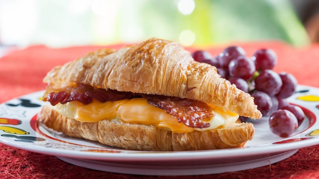 Sándwich de croissant con huevos revueltos, queso y tocineta en un plato con uvas