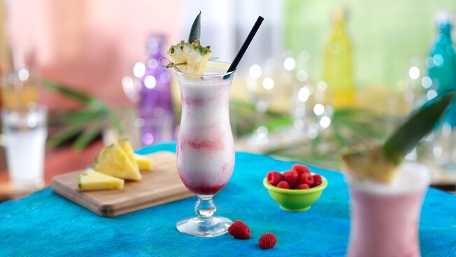 Tall glass of Piña CoLAVA cocktail with straw and pineapple spear and bowl of raspberries on table