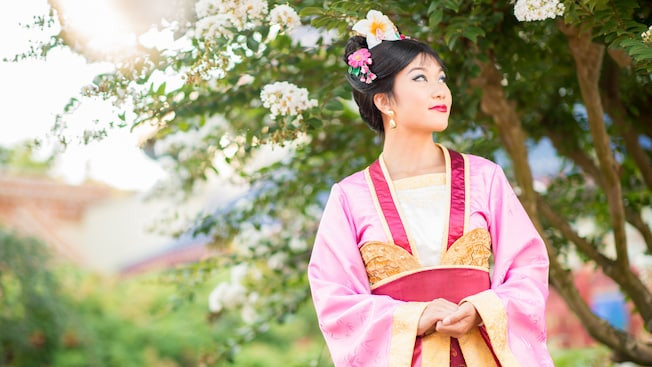 Mulan wears a colorful Chinese robe and flowers in her hair as she gazes into the distance