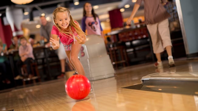 A smiling young girl bowls with both hands while her family watches in the background at Splitsville Luxury Lanes in Downtown Disney Area
