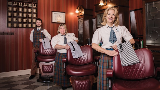 2 women and a man pose with aprons by barber shop chairs