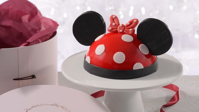 An Amorette’s Patisserie Minnie Mouse dome cake on a pedestal next to its hat box packaging
