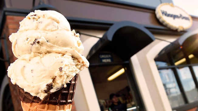 A chocolate dipped cone with 3 scoops of vanilla chocolate chip ice cream is held in front of the exterior of Haagen Dazs