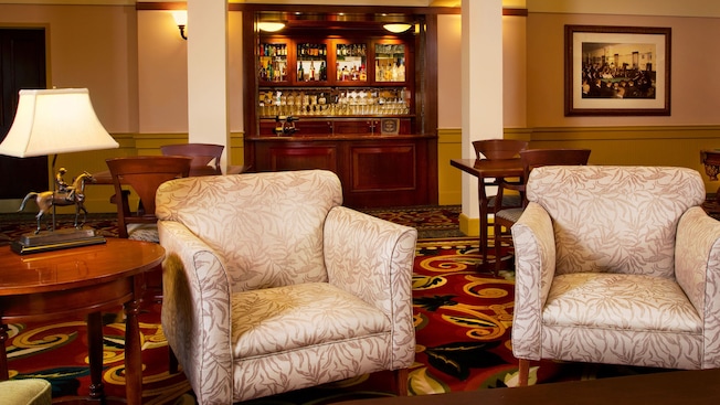 Un salón alfombrado con varias mesas de juego de madera y sillas, un sillón tapizado, una lámpara de mesa con un caballo de bronce, una mesa auxiliar de madera y un bar empotrado con vino, licores y otras bebidas