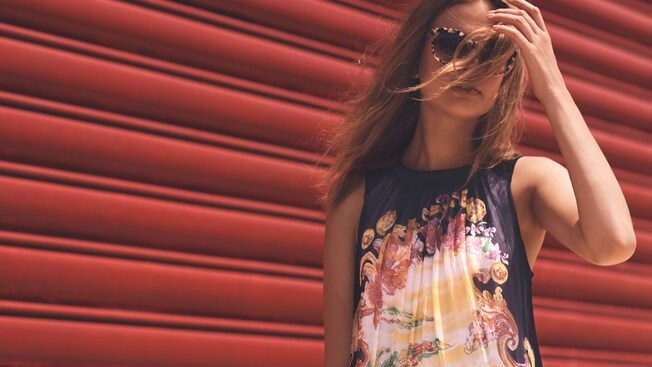 A woman wearing sunglasses and a sleeveless top from Anthropologie brushes a section of hair away from her face