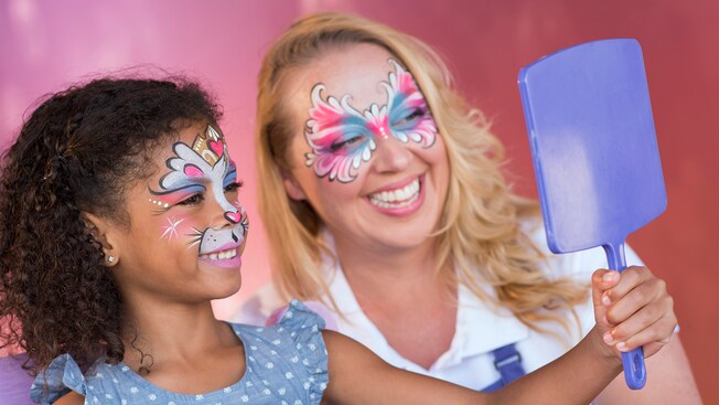 Una niña sostiene un espejo para observar su maquillaje en el quiosco de Maquillaje Artístico de Enjoy Your Face en Disney Springs mientras la artista que la maquilló la observa
