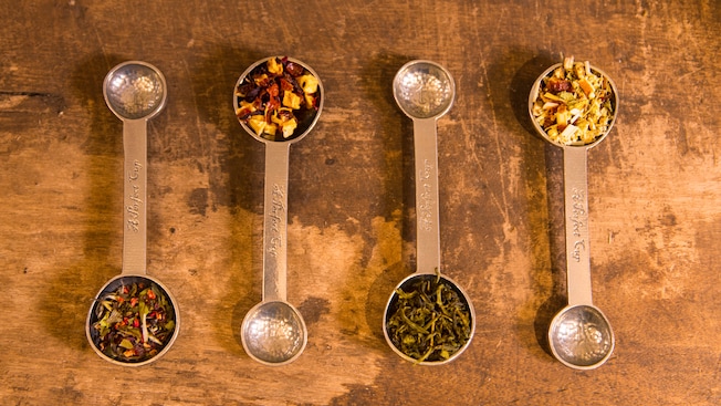 Four double ended measuring spoons laid side by side each containing a different loose leaf tea in the larger end