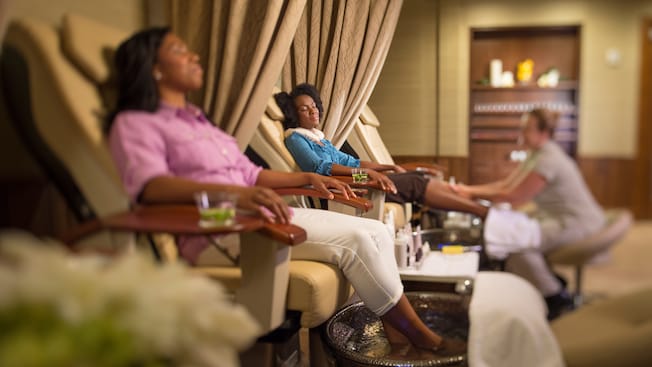 A mother and her grown daughter relax in reclining chairs as a manicurist gives the daughter a pedicure