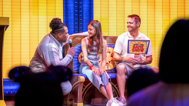 Uma garota, um homem e um apresentador em uma atividade de disputa de prêmios no lounge Saga a bordo do navio Disney Destiny
