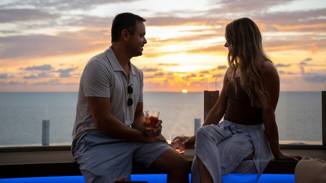 Um casal apreciando bebidas e o pôr do sol em um banco ao ar livre no Infinity Bar, no Disney Adventure