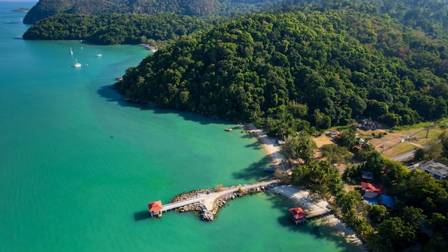 A densely forested tropical island surrounded by water, with a jetty extending into the sea and traditional village buildings along the shoreline A densely forested tropical island surrounded by water, with a jetty extending into the sea and traditional village buildings along the shoreline