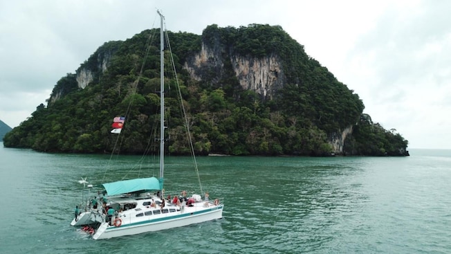 A catamaran cruising on the water near a large, forested cliff A catamaran cruising on the water near a large, forested cliff