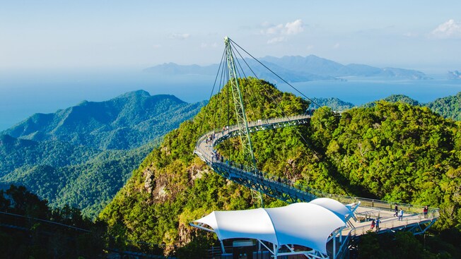 A curved pedestrian suspension bridge arcs across a lush mountain ridge above the Langkawi jungle