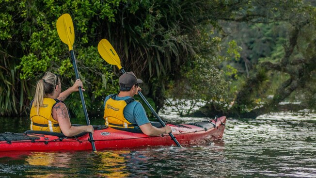 Two Guests with life jackets paddling a double kayak along a forested shore