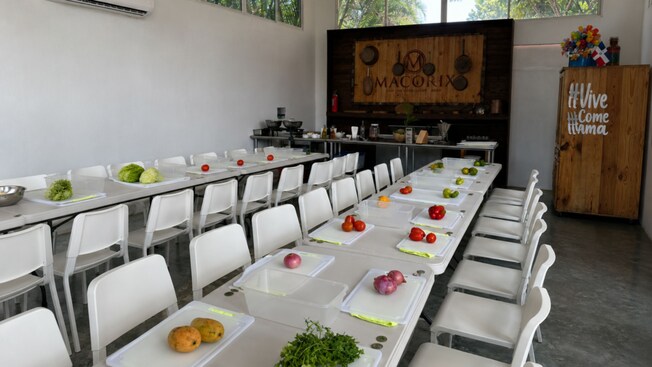 A cooking classroom featuring fresh produce sitting on the cutting board in front of each chair at long tables