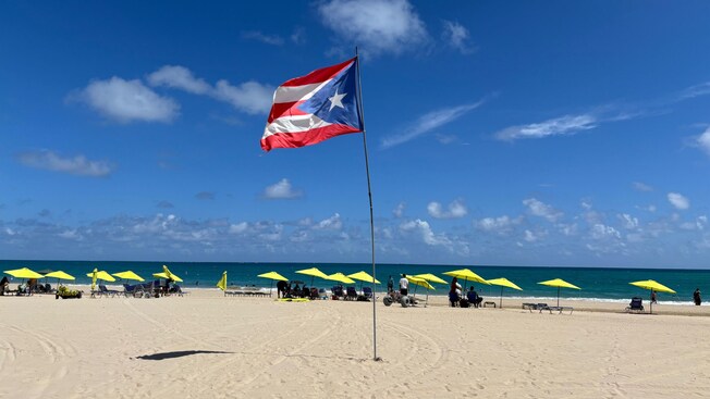 A Puerto Rican territorial flag and shaded outdoor furniture on a sandy beach in San Juan