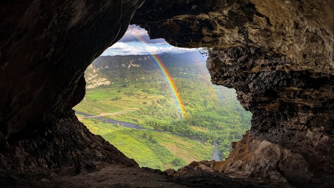 A rainbow over a lush valley and river, framed by the opening of a cave