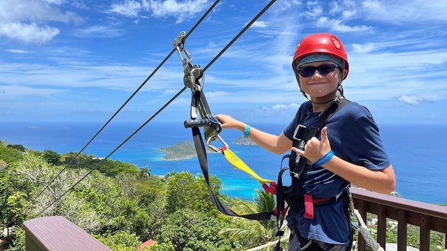 A boy posing on a platform while tethered to a zip line pulley with the coastline behind him A boy posing on a platform while tethered to a zip line pulley with the coastline behind him