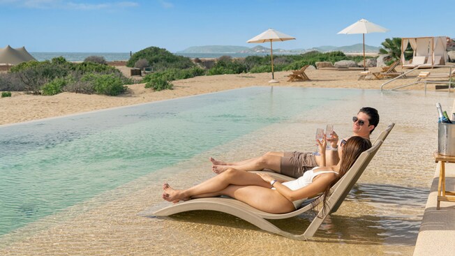 A couple in chaise lounges clinking glasses while relaxing in a shallow pool right on the beach