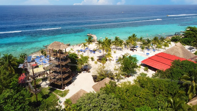 An aerial view of Islands Beach Club in Cozumel, Mexico featuring a beach, palm trees, lounge chairs, a swimming pool and a wooden tower with zip lines An aerial view of Islands Beach Club in Cozumel, Mexico featuring a beach, palm trees, lounge chairs, a swimming pool and a wooden tower with zip lines