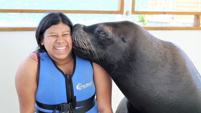 A smiling woman in a life jacket being kissed by a sea lion