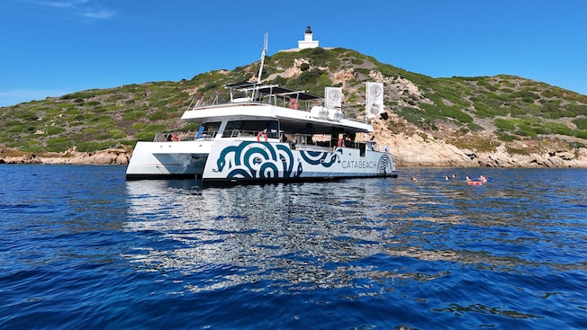A catamaran anchored in calm waters near a hillside with a lighthouse in Ajaccio, Corsica A catamaran anchored in calm waters near a hillside with a lighthouse in Ajaccio, Corsica
