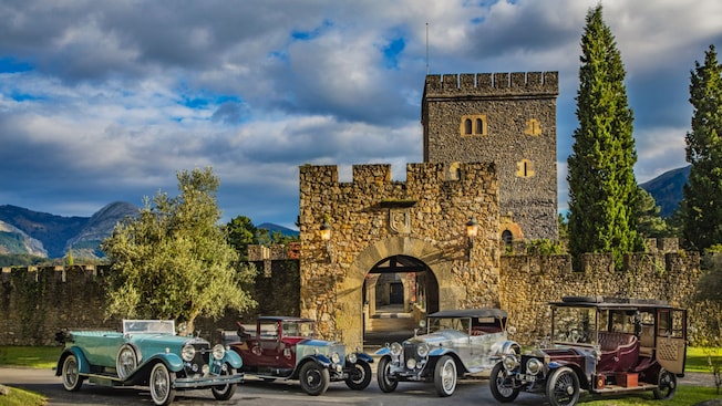 Cuatro autos de lujo clásicos estacionados frente a un castillo de piedra histórico con una torre, una entrada arqueada y vegetación circundante Cuatro autos de lujo clásicos estacionados frente a un castillo de piedra histórico con una torre, una entrada arqueada y vegetación circundante