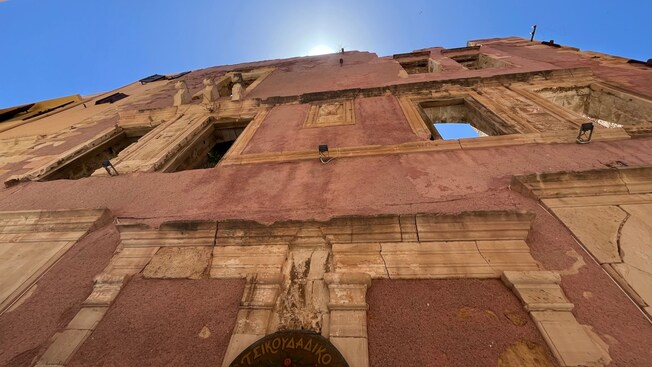 The upper facade of a crumbling building with open windows framed by narrow alley walls