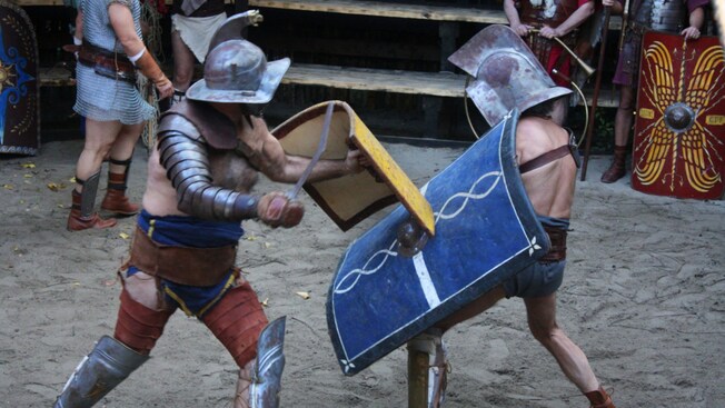 Two gladiators in armor and shields fighting in a sandy arena with spectators nearby