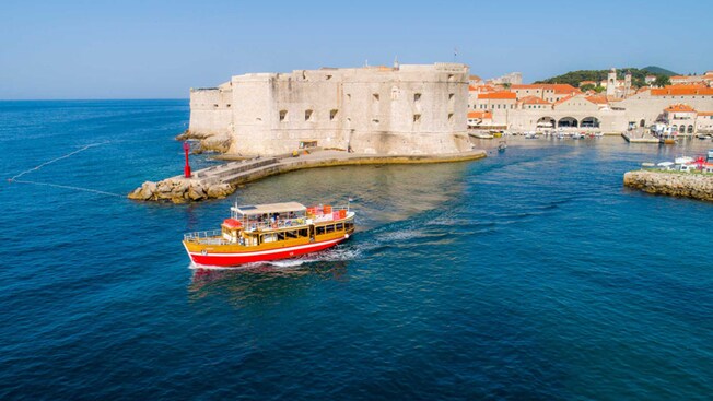 Un barco turístico de madera navegando frente a la entrada de la fortaleza de piedra del Puerto Viejo de Dubrovnik