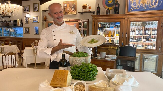 Chef en una mesa de un restaurante mostrando ingredientes como queso, albahaca, piñones y aceite de oliva, con un plato de pasta