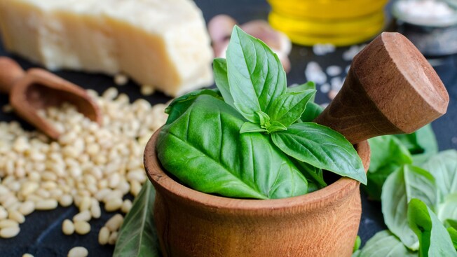 A wooden pestle and mortar filled with fresh basil on a table next to pine nuts and parmesan cheese