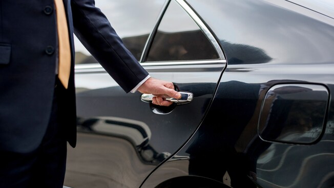A professional driver in a suit opening the rear door of a luxury sedan