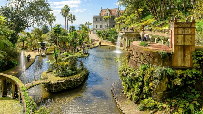 El jardín tropical Monte Palace en Madeira, Portugal, cuenta con un lago con pequeñas islas y una cascada, rodeada por una frondosa vegetación y un camino que conduce a un gran edificio