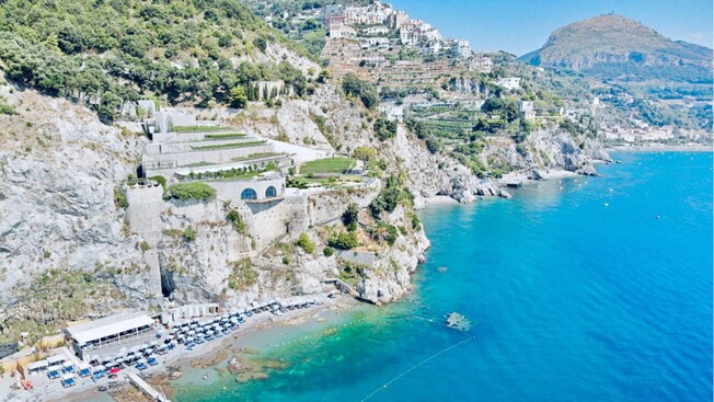 A beach with designated swimming areas and lounge chairs below a series of terraced gardens on a rocky cliff