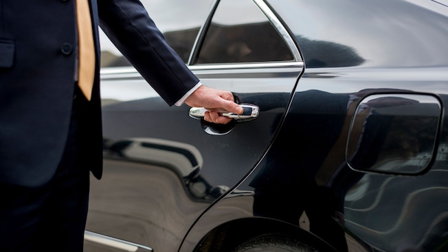 A man wearing a suit while holding the handle of a car door A man wearing a suit while holding the handle of a car door