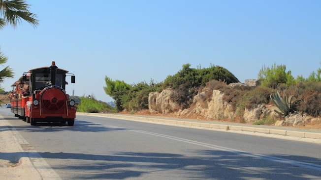 A tourist trolley train traveling along a paved road with rocky terrain and greenery A tourist trolley train traveling along a paved road with rocky terrain and greenery