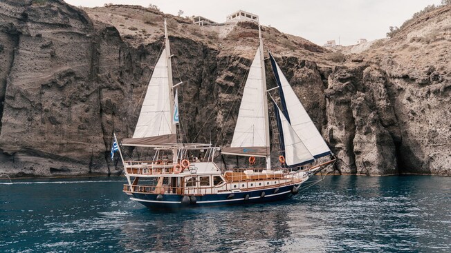 The Adonis luxury schooner sailing on the water near a cliff in Santorini, Greece