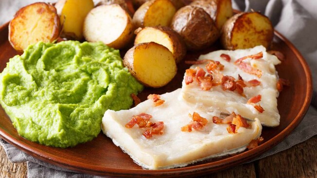 A plate filled with traditional Norwegian food including mashed peas, roasted potatoes and fish topped with pieces of bacon