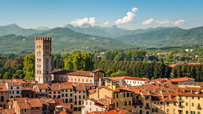 A scenic view featuring historic buildings with terracotta rooftops, a tall bell tower and a backdrop of mountains