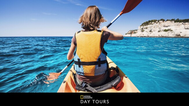 Un Visitante con chaleco salvavidas paseando en kayak en aguas abiertas con acantilados a la distancia y las palabras “National Geographic Day Tours”