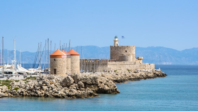 A stone fortress with a lighthouse and 2 windmills on a rocky pier, surrounded by sailboats and calm water A stone fortress with a lighthouse and 2 windmills on a rocky pier, surrounded by sailboats and calm water