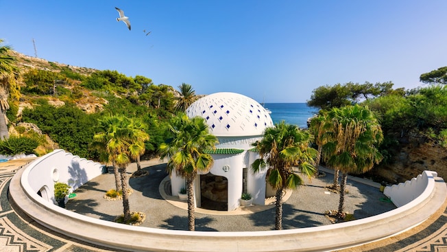 A domed building surrounded by palm trees and a circular decorative courtyard, overlooking the sea A domed building surrounded by palm trees and a circular decorative courtyard, overlooking the sea