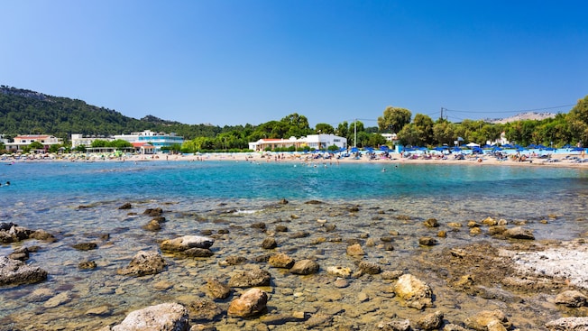 A rocky shoreline in Rhodes, Greece, featuring clear water, lounge chairs and coastal buildings