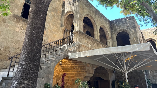 Historic stone building in Rhodes featuring arched walkways, a staircase and leafy trees shading a courtyard Historic stone building in Rhodes featuring arched walkways, a staircase and leafy trees shading a courtyard