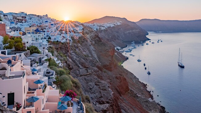 Traditional Greek buildings cascading down the cliffside with sailboats in the water below